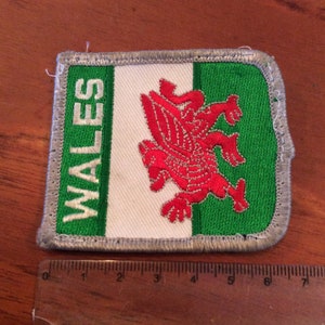 May include: A green, white, and red embroidered patch with the word "WALES" and a red dragon on a white background.