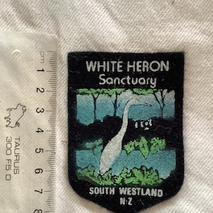Peut inclure: Un écusson noir et blanc avec un héron blanc debout dans un étang bleu avec des roseaux verts. L'écusson porte les inscriptions "White Heron Sanctuary" et "South Westland N.Z."