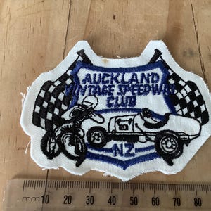 Puede incluir: Parche bordado con el texto "Auckland Vintage Speedway Club" en azul y blanco. El parche muestra una motocicleta y un coche de carreras, con banderas a cuadros. También son visibles las letras "NZ".