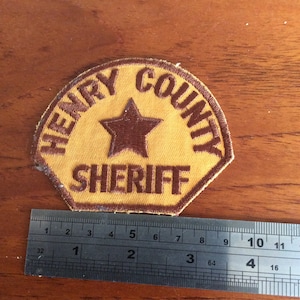 Könnte beinhalten: Ein gestickter Aufnäher in Braun und Gelb mit dem Text "HENRY COUNTY SHERIFF" und einem braunen fünfzackigen Stern in der Mitte. Der Aufnäher wird mit einem Lineal gemessen und ist etwa 6,5 cm breit.