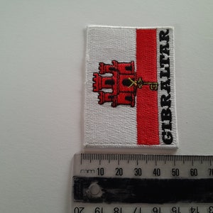 Può includere: Una toppa ricamata bianca e rossa con lo stemma di Gibilterra e la parola "GIBRALTAR" scritta verticalmente sul lato destro.