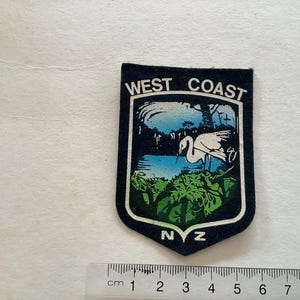 May include: A blue and white embroidered patch with the text "West Coast NZ" and a white bird in a blue lake surrounded by green foliage.