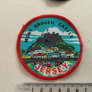 May include: A round, red-bordered patch with a blue and green image of Mont Orgueil Castle in Jersey. The text "Mont Orgueil Castle Jersey" is printed in black around the image.