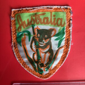 May include: A vintage patch with an orange border and the word "Australia" in green script. The patch features a cartoon koala bear clinging to a bamboo stalk. The koala is brown and white, and the bamboo is green and orange.