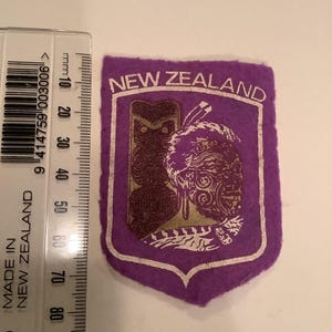 May include: A purple felt patch with the words "NEW ZEALAND" in white. The patch features two brown, stylized Maori carvings within a white border. A ruler is placed next to the patch, showing measurements in millimeters.