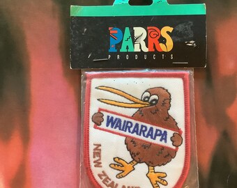 Op de afbeelding: Een geborduurde patch met een kiwi die een bord vasthoudt met de tekst "Wairarapa, Nieuw-Zeeland". De patch is bruin en wit en verpakt in een doorzichtige plastic zak met een rood, wit en blauw label met de tekst "Parrs Products".