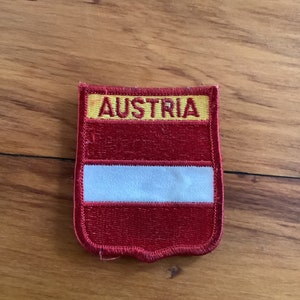 May include: A red and yellow embroidered patch with the word "AUSTRIA" in yellow lettering and a white stripe in the center.
