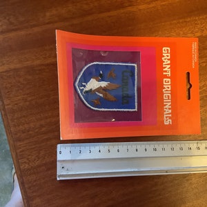 May include: A blue and white embroidered patch featuring a Canadian goose and the word "Canada" on a shield. The patch is packaged in a plastic sleeve with an orange background and the text "GRANT ORIGINALS" printed on the side.
