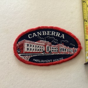May include: Oval patch featuring the text "CANBERRA" above an illustration of Parliament House. The patch is navy blue with a red border and the text "PARLIAMENT HOUSE" below the building. The patch measures approximately 3 inches in length.