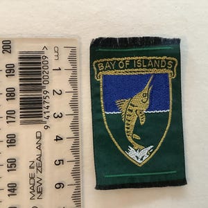 May include: A green and gold embroidered patch with the text "BAY OF ISLANDS" at the top. The patch features a blue background with a marlin fish design. The patch is approximately 6 cm in length.