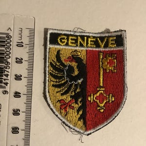 May include: Embroidered patch featuring the word "GENEVE" above a shield design. The shield is divided into yellow and red sections, with a black eagle and a gold key. The patch is approximately 2.5 inches wide.