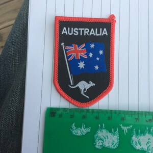 May include: An embroidered patch with the word "AUSTRALIA" above the Australian flag and a kangaroo. The patch is black with a red border. The patch is approximately 7.6 cm tall and 5 cm wide.