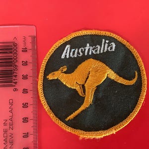 Può includere: Toppa rotonda con la scritta "Australia" in bianco sopra una silhouette di canguro dorato su sfondo nero. La toppa ha un bordo dorato. Un righello rosso con segni neri e testo bianco è nell'immagine.