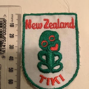 May include: A white, shield-shaped patch with a green border. The patch features the words "New Zealand" in red, and a green Tiki design. The word "TIKI" is written in red at the bottom of the patch.