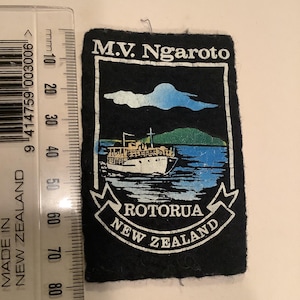 Op de afbeelding: Zwarte patch met de tekst "M.V. Ngaroto ROTORUA NEW ZEALAND". De patch toont een wit schip op blauw water, met groen land en een blauwe lucht. De patch is ongeveer 7,6 cm breed.