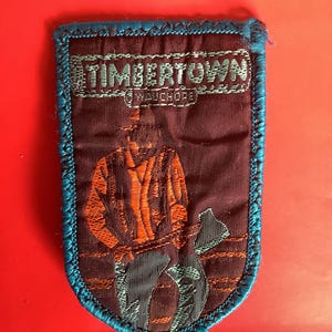 Puede incluir: Un parche rectangular con borde azul y fondo granate. El parche presenta el texto "TIMBERTOWN" sobre una ilustración de un leñador sosteniendo un hacha. El leñador lleva una camisa naranja y pantalones azules.