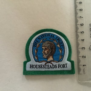 May include: A green felt patch with white embroidered text that reads "The National Trust" and "Housesteads Fort". The patch features a blue circle with a brown image of a person's head inside a wreath.