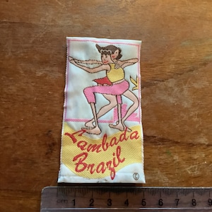 Puede incluir: Un parche de tela blanca con un diseño bordado en amarillo, rojo y rosa de dos mujeres bailando. El texto "Lambada Brazil" está bordado en rojo y rosa en el parche.