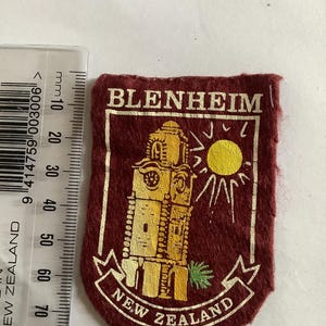 May include: A maroon fabric patch with a gold tower illustration, a sun, and the words "BLENHEIM" and "NEW ZEALAND". The patch is next to a ruler showing measurements in millimeters. The patch is approximately 6 cm in length.