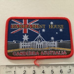 以下が含まれることがあります： オーストラリアのキャンベラにある国会議事堂を描いた刺繍パッチ。オーストラリアの国旗、テキスト「PARLIAMENT HOUSE」と「CANBERRA AUSTRALIA」が赤い縁取りのある青い背景にデザインされています。パッチは長方形です。