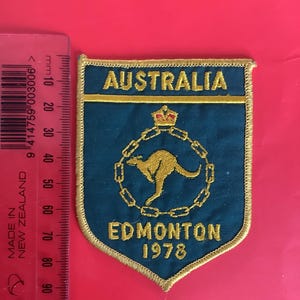 Può includere: Toppa blu a forma di scudo con ricamo dorato. La toppa riporta la scritta "AUSTRALIA" in alto e "EDMONTON 1978" in basso. Un canguro dorato è al centro, circondato da una catena.