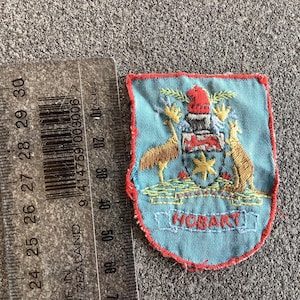 May include: Embroidered patch with a light blue background and a red border. The patch features a coat of arms with a kangaroo and emu on either side. The word "HOBART" is embroidered at the bottom. A ruler is next to the patch.