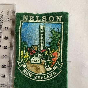 Pode incluir: Remendo de feltro verde com a palavra "NELSON" no topo e "NEW ZEALAND" em uma faixa na parte inferior. O remendo apresenta uma ilustração colorida de uma torre, edifícios e paisagem. Uma régua está no lado esquerdo.