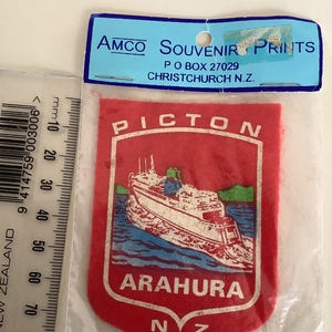 Op de afbeelding: Een rode souvenirpatch met een witte en blauwe illustratie van een veerboot op het water, met de woorden "PICTON" en "ARAHURA" in het wit. De patch zit in een doorzichtige plastic hoes met een blauwe header "AMCO SOUVENIR PRINTS".