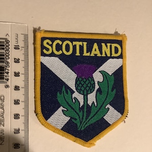 Pode incluir: Um patch em forma de escudo com a palavra "SCOTLAND" em amarelo sobre um fundo azul marinho. Apresenta um desenho de cruz branco e azul e um cardo verde com uma flor roxa. O patch tem uma borda amarela.