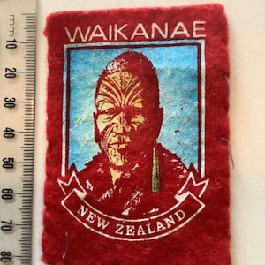 May include: A rectangular red felt patch with the text "WAIKANAE" at the top and "NEW ZEALAND" on a banner at the bottom. The patch features a portrait of a person with traditional facial markings, set against a blue background.