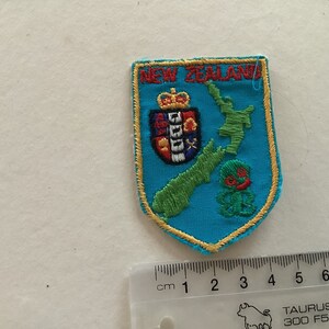 May include: A blue embroidered patch with the words "New Zealand" in yellow lettering. The patch features a green embroidered outline of the country's North and South Islands, a red and gold embroidered coat of arms, and a green embroidered kiwi bird.
