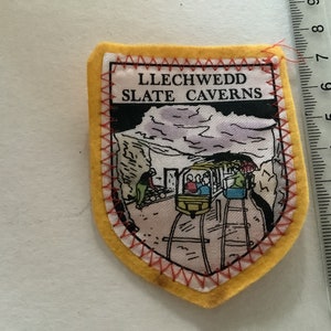 Puede incluir: Un parche de fieltro amarillo con costuras rojas. El parche presenta una ilustración en blanco y negro de un tren entrando en una cueva con el texto "LLECHWEDD SLATE CAVERNS".