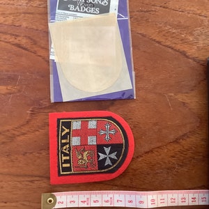Può includere: Una toppa ricamata rossa, bianca e nera con la parola "ITALIA" e uno stemma con un leone, una croce e un motivo a scacchi. La toppa è su una superficie di legno.