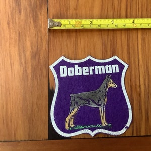 May include: Purple felt patch with a white border and the word "Doberman" in white lettering. A black and tan Doberman Pinscher is depicted in the center of the patch.