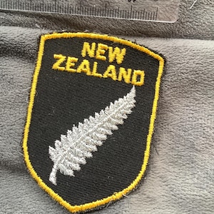 May include: A black and gold embroidered patch with the words "NEW ZEALAND" in gold lettering. The patch features a silver fern design. The patch is shield-shaped.