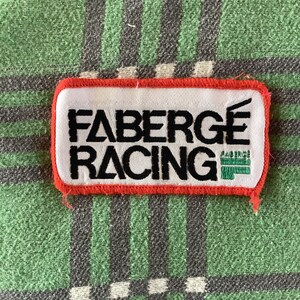 Könnte beinhalten: Ein weißer und roter Stoff-Patch mit dem Text "FABERGÉ RACING" in schwarzen Stickbuchstaben.