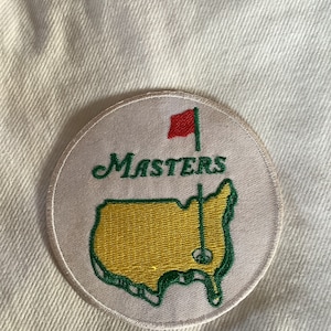 Puede incluir: Un parche circular con la palabra "Masters" en escritura verde sobre un mapa verde y amarillo de los Estados Unidos con una bandera de golf roja. El parche está sobre una tela color crema.