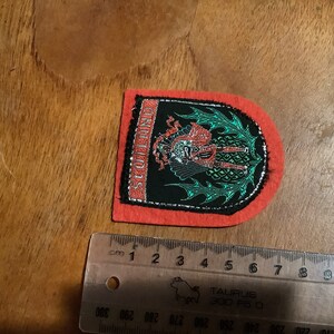 May include: A red felt patch with a black and green embroidered design of a dragon with the word "Scotland" written vertically on the left side.