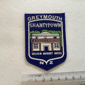 May include: A blue and white patch with the text "GREYMOUTH SHANTYTOWN GOLDEN NUGGET HOTEL NZ". The patch features a cartoon illustration of a building with a brown roof and white walls.
