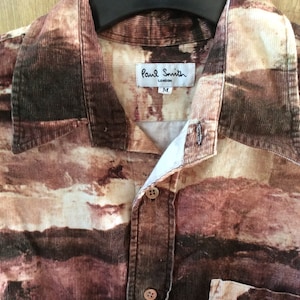 May include: A brown, beige, and white corduroy shirt with a landscape print. The shirt has a button-up front and a patch pocket on the left side. The label reads "Paul Smith London" and "M".