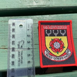 May include: A red, black and yellow embroidered patch with the text "DERBYSHIRE" and a crest featuring a red rose and three stag heads. The text "BENE CONSULENDOS" is written in a silver-coloured script around the bottom of the crest.