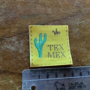 以下が含まれることがあります： 黄色の布のパッチに、緑色のサボテンと茶色の文字で「TEX MEX」と書かれています。右上の隅には、馬に乗った人の小さな茶色のシルエットがあります。