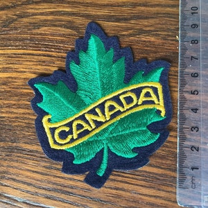 Puede incluir: Una hoja de arce bordada en fieltro verde con una pancarta de fieltro amarillo que dice "CANADA". El parche está sobre una superficie de madera marrón oscura.