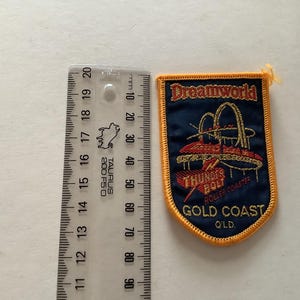 May include: A navy blue patch with gold embroidered text and a roller coaster design. The patch reads "Dreamworld Thunder Bolt Roller Coaster Gold Coast QLD". The patch is approximately 3 inches tall.