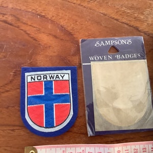 May include: A blue and red woven patch featuring the word "NORWAY" and the Norwegian flag. The patch is packaged in a clear plastic bag with a white label that reads "SAMPSON'S WOVEN BADGES".