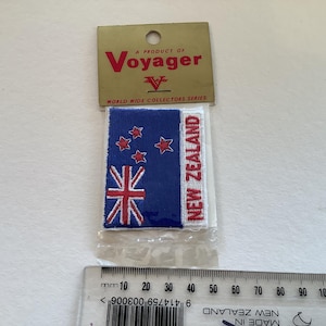 Può includere: Una carta bianca con una toppa ricamata blu e rossa della bandiera della Nuova Zelanda. La carta dice "A Product of Voyager" e "World Wide Collectors Series".