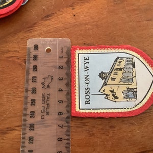 以下が含まれることがあります： 「ROSS-ON-WYE」という文字と建物の絵が描かれた赤、白、青の刺繍パッチ。定規で測定したところ、幅は約6cmです。