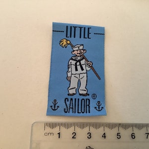 May include: A light blue fabric label featuring the words "LITTLE SAILOR" and a cartoon sailor holding a mop. The label includes two anchors and a registered trademark symbol.