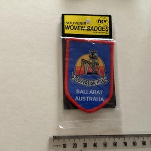 Könnte beinhalten: Ein gewebtes Souvenir-Abzeichen in Blau und Rot mit dem Text "SOVEREIGN HILL BALLARAT AUSTRALIA". Das Abzeichen zeigt ein Bergwerksgebäude und ist in einer transparenten Kunststoffhülle verpackt.
