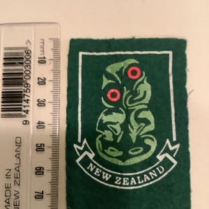 May include: A green felt patch with a white border and a stylized green and red design. The patch features the words "NEW ZEALAND" in white on a banner. The patch is approximately 2.5 inches tall.